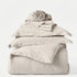 Stack of beige bedding including a duvet, pillowcases, and a blanket on a white background