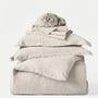 Stack of beige bedding including a duvet, pillowcases, and a blanket on a white background